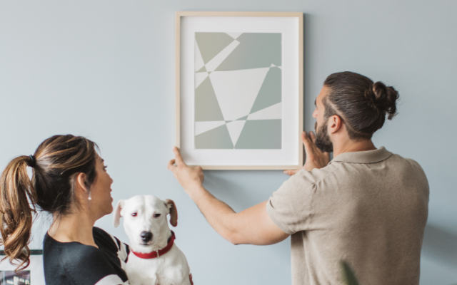 A couple with a dog hanging a picture on a wall
